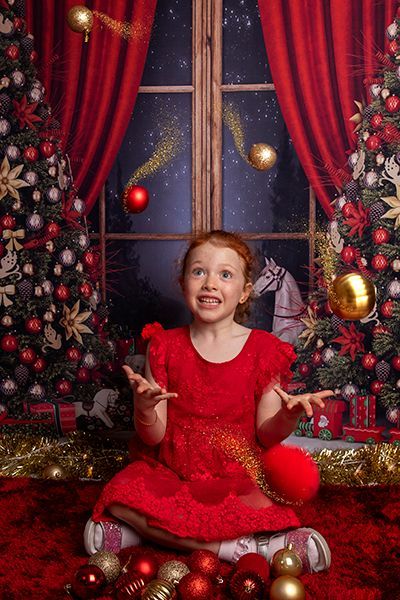 A little girl in a red dress is sitting on the floor in front of a christmas tree.