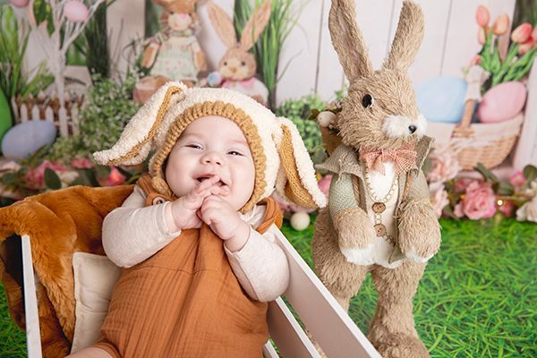 A baby wearing a bunny hat is sitting in a wooden crate next to a stuffed bunny.