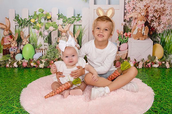 A boy and a baby are sitting on a pink rug holding carrots.