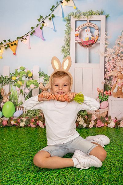 A little boy wearing bunny ears is sitting on the grass holding a carrot in his mouth.