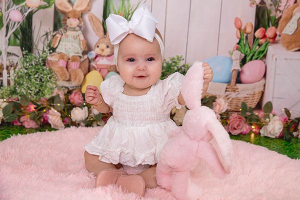 A baby in a white dress is sitting on a pink blanket holding a stuffed bunny.