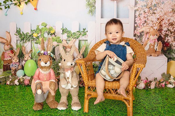 A baby is sitting in a wicker chair next to stuffed rabbits.