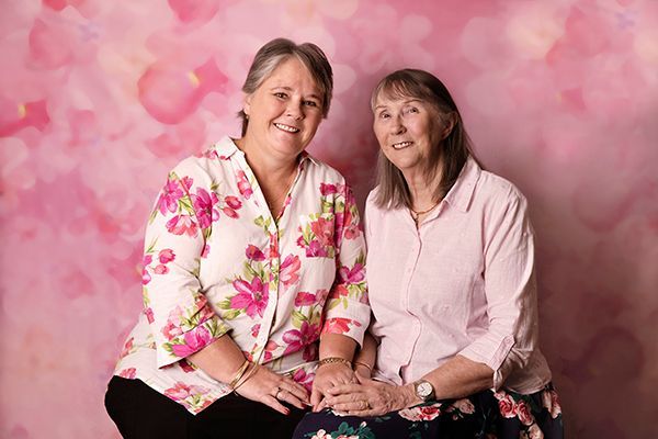 Two women are sitting next to each other in front of a pink background.