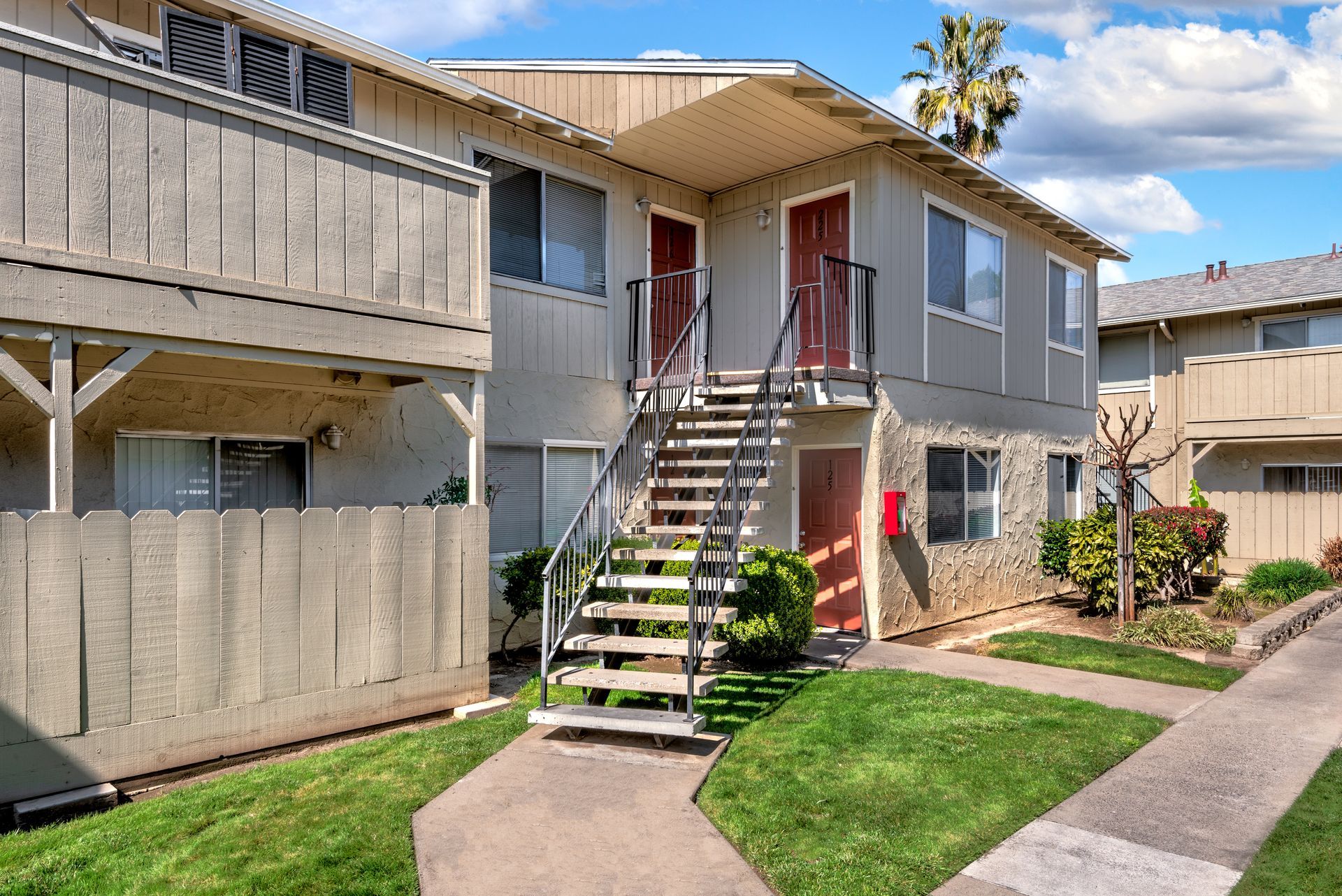 Two-story apartment building with exterior staircases, red doors, and green grass on a sunny day.