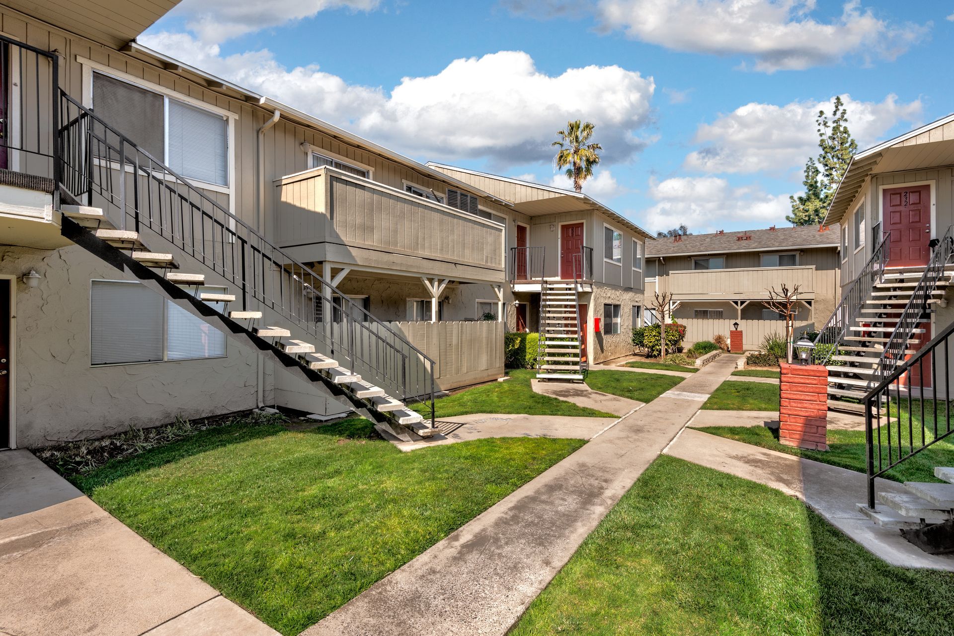 Apartment complex with beige buildings, exterior stairs, grass, walkways, and a blue sky.