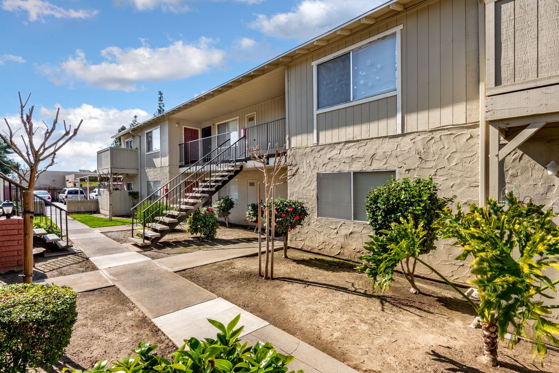 Two-story beige apartment building with outdoor staircases, pathway, and greenery under a cloudy sky.