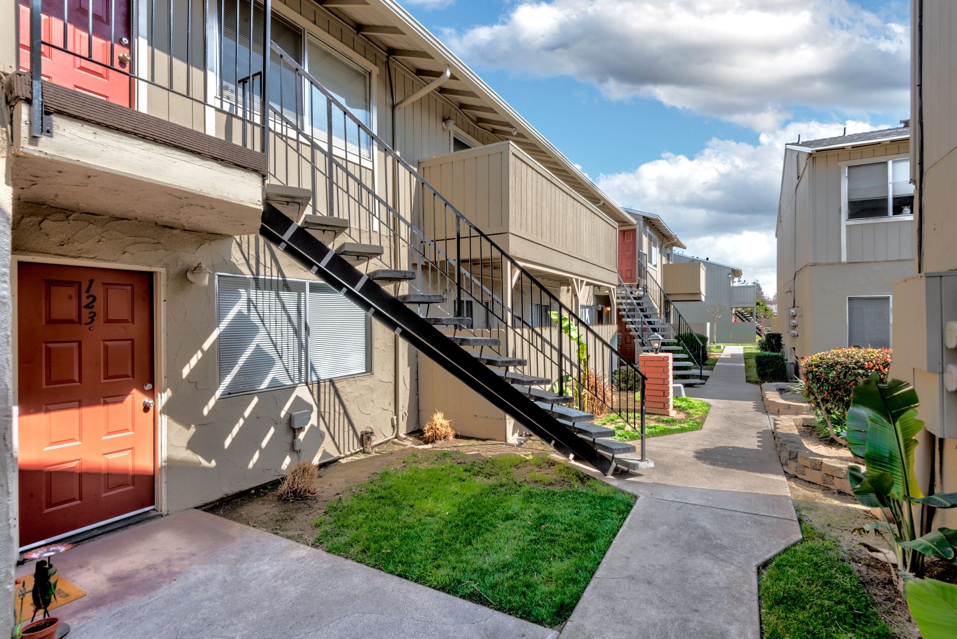 Exterior of a two-story apartment complex with stairs, a walkway, and small lawns; sunny day.