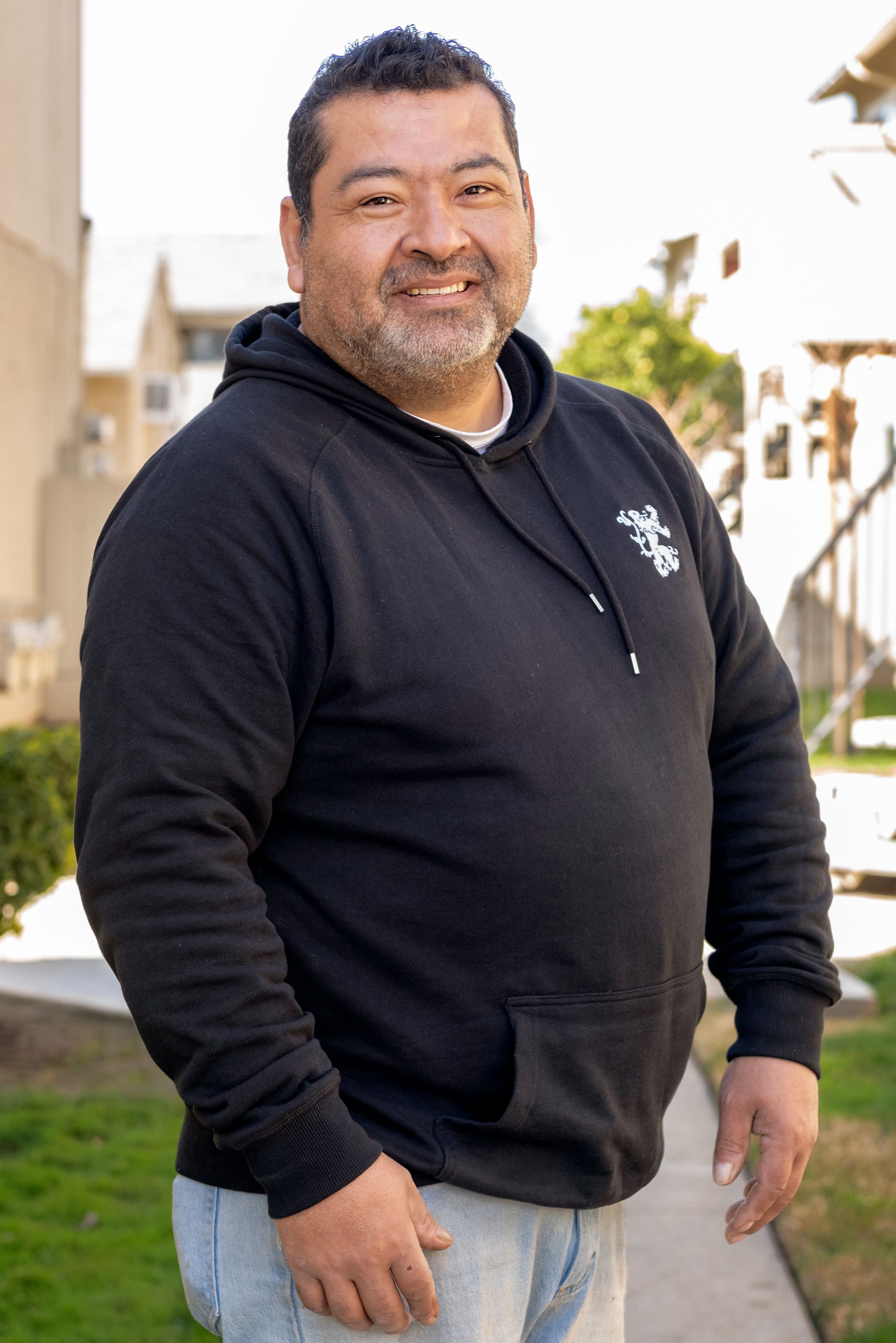 Man smiling, wearing a black hoodie and standing outside apartment buildings.