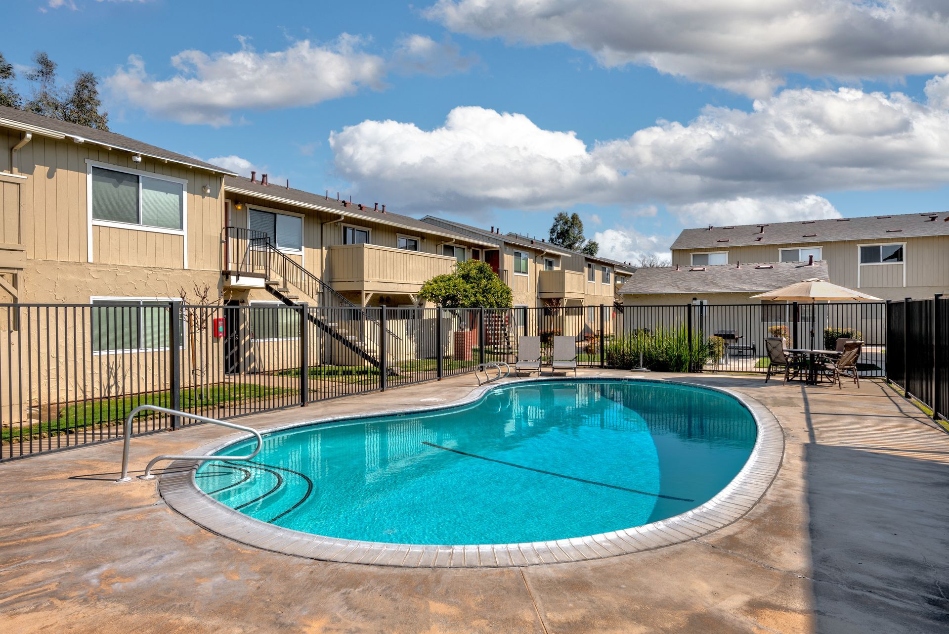 Apartment complex with a pool. Beige buildings, black fence, blue pool, sunny day.