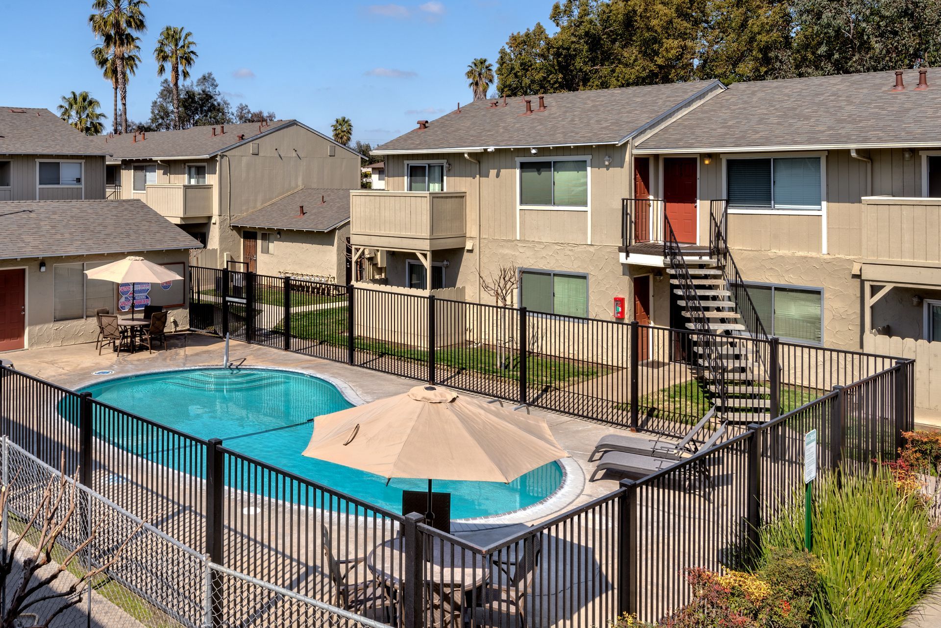 Apartment complex with swimming pool, beige buildings, black fence, tan umbrellas, and palm trees in background.