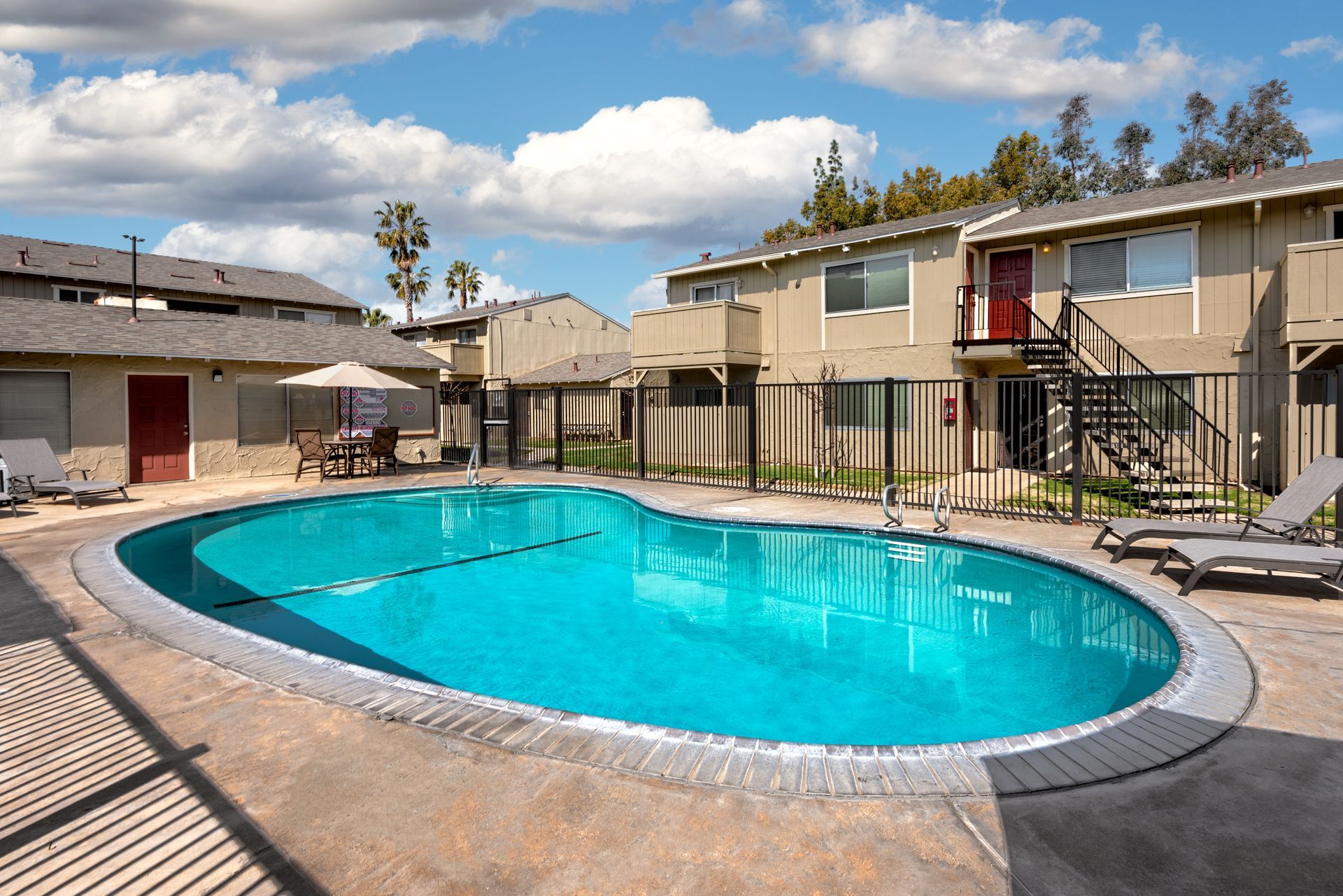 Swimming pool surrounded by apartments under a blue sky. Lounge chairs are visible.