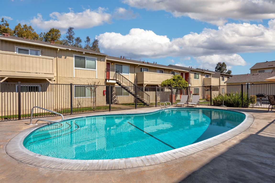 Apartment complex with pool; beige buildings, blue water, fence, cloudy sky.