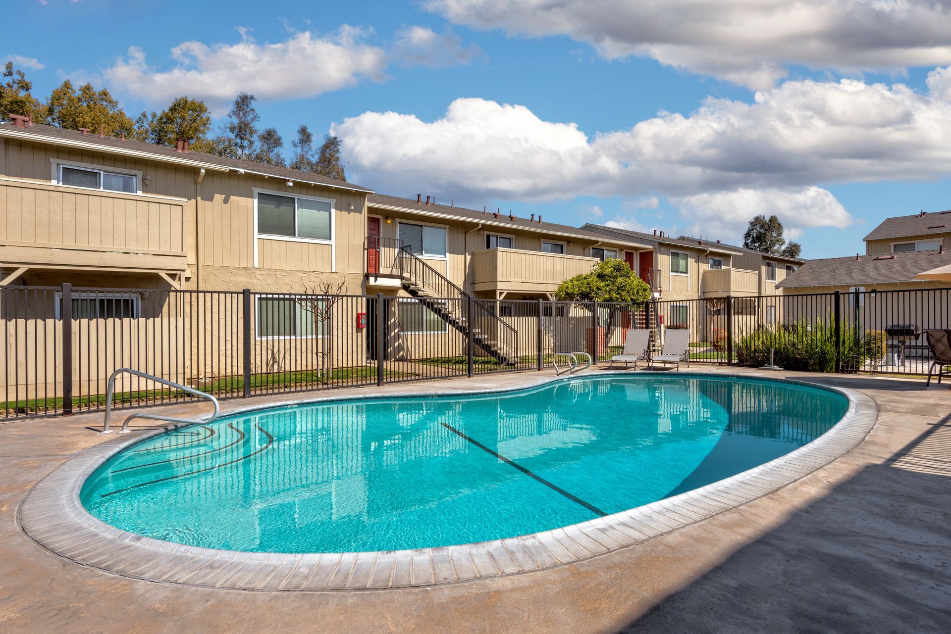 Apartment complex with pool; beige buildings, blue water, fence, cloudy sky.