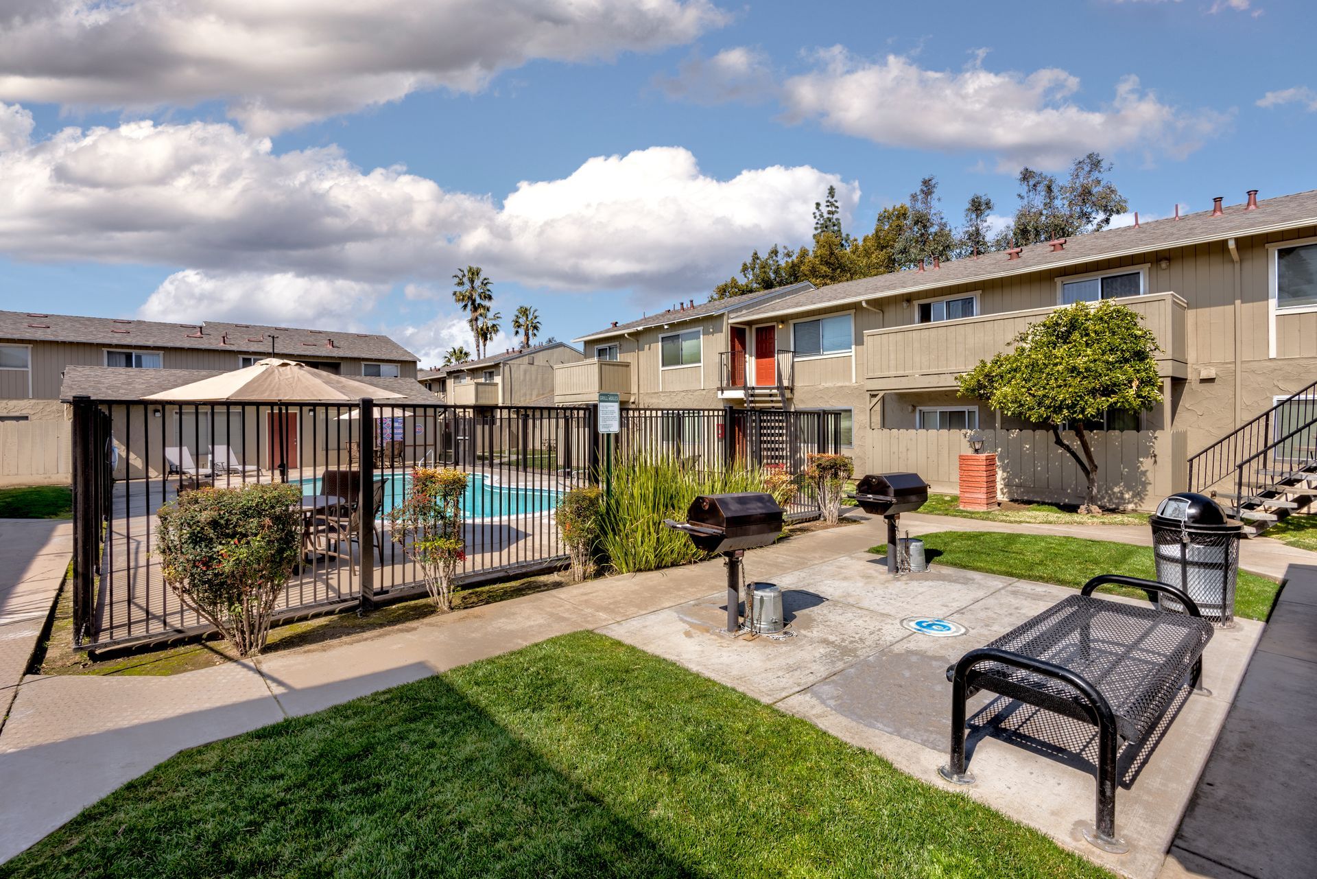 Apartment complex courtyard with pool, grills, and seating under a partly cloudy sky.