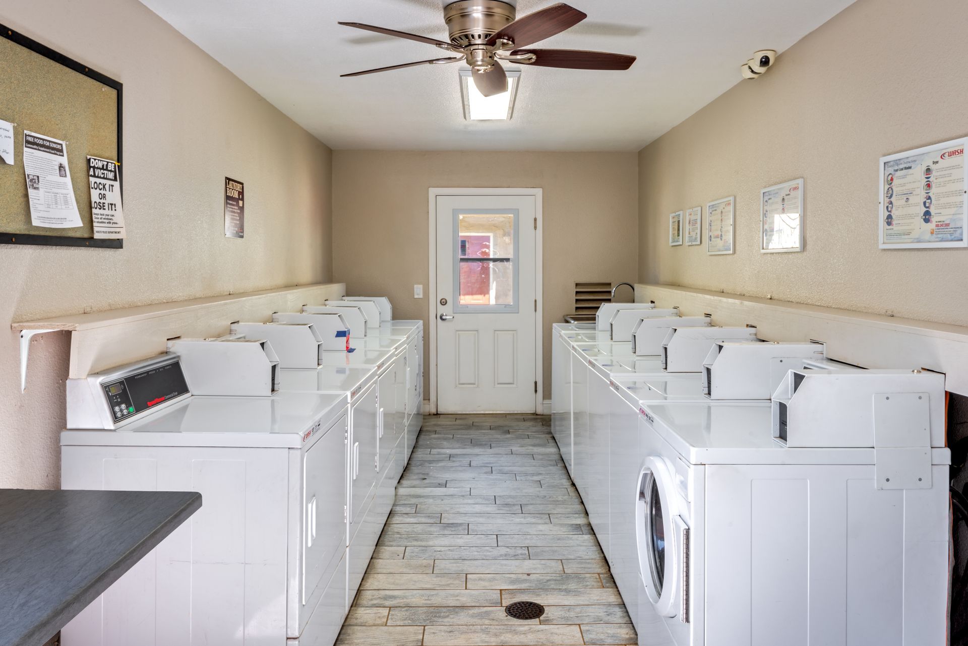 A laundry room with rows of white washing machines and dryers, neutral walls, and a ceiling fan.