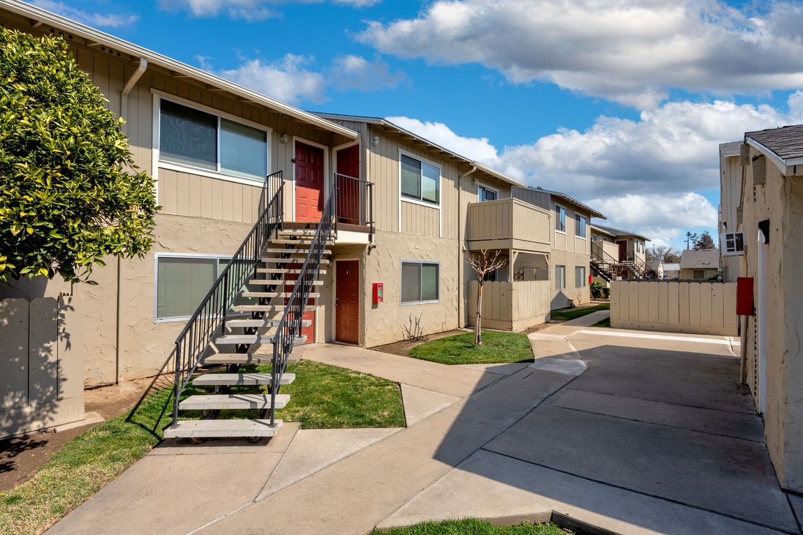 Apartment complex exterior with tan buildings, stairs, and green grass under a cloudy blue sky.