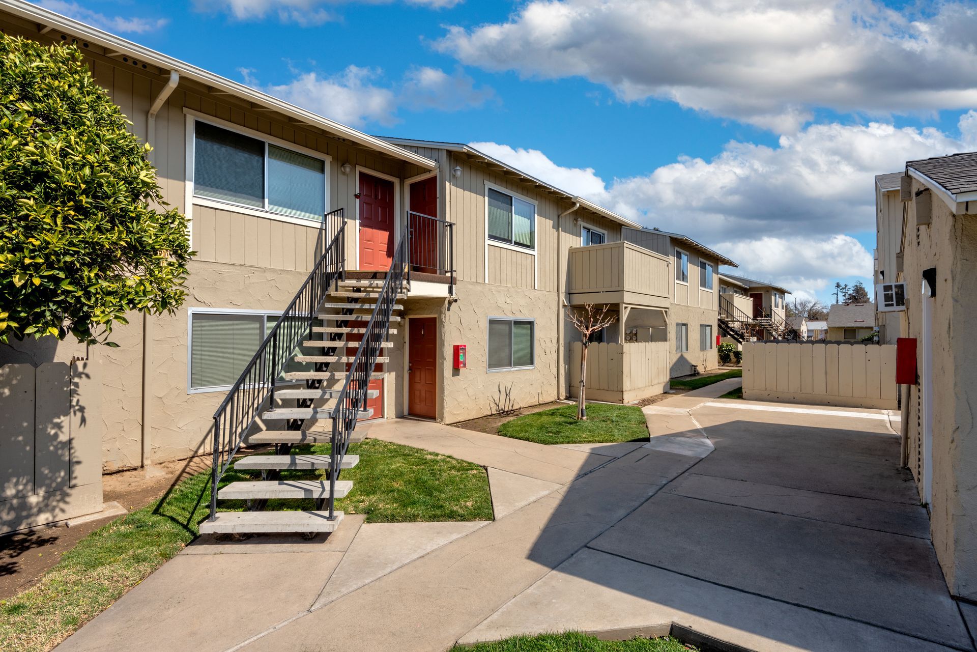 Apartment complex exterior with tan buildings, stairs, and green grass under a cloudy blue sky.