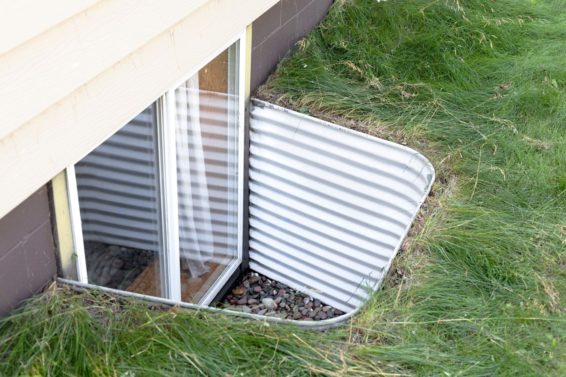 Basement window well with corrugated metal covering beside a house and grass yard