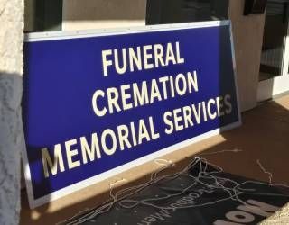 Blue sign reading “FUNERAL CREMATION MEMORIAL SERVICES” leaning against a wall, with cords on the floor.