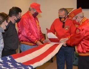 Several men in red jackets and masks fold a large American flag indoors.