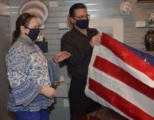 Two masked people indoors hold a red-and-white striped flag in a room with shelves.