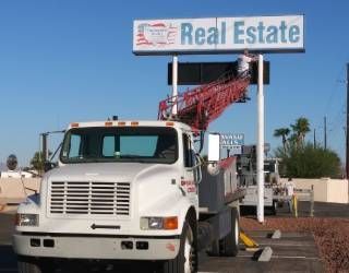 White truck beside a Real Estate sign with a red crane boom in a parking lot
