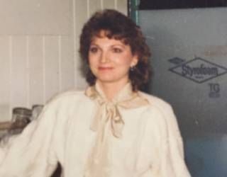 Woman in a cream blouse with a scarf, standing indoors beside a refrigerator.