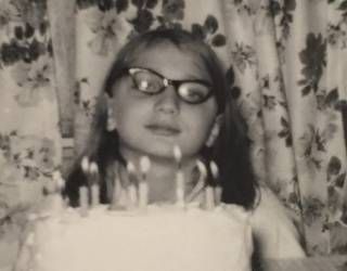 Person wearing glasses behind a birthday cake with lit candles, in front of a floral curtain