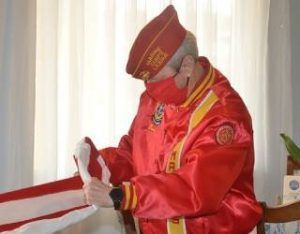 Person in red ceremonial uniform and cap, holding a folded red-and-white fabric indoors by a bright window