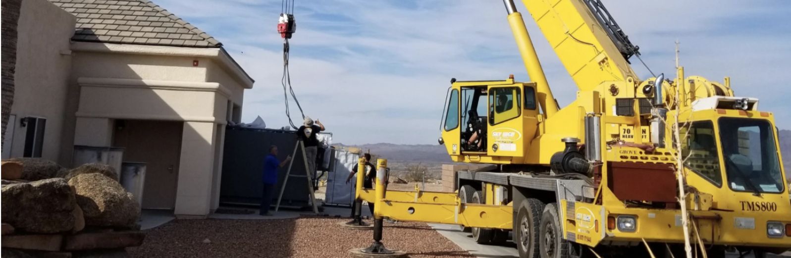 Yellow crane truck parked beside a house, lifting a large load hook in a residential driveway.