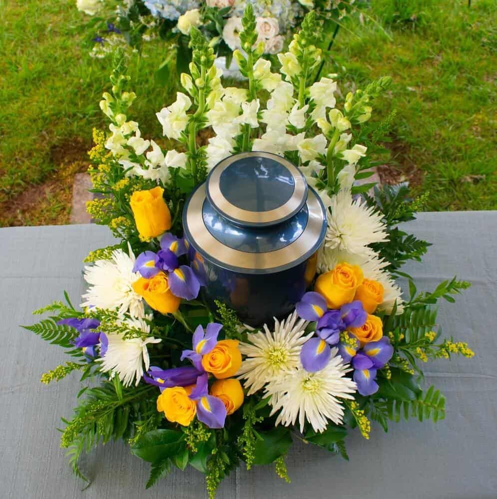 Dark grey urn with bronze colored band with floral arrangement surrounding it sitting on concrete with grass in the background. 
