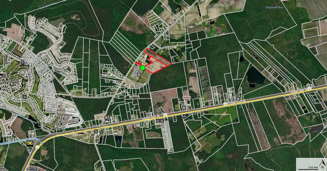 Aerial view of green and brown fields, roads, and a structure outlined in red with a green dot.