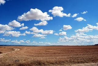 A dirt road going through a dry field with clouds in the sky