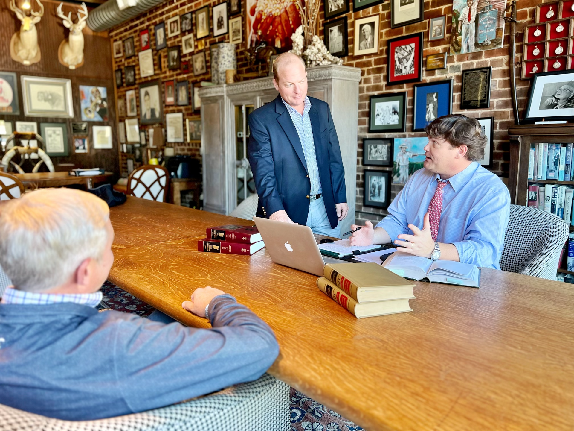 Three men in a room with a wooden table, discussing with a laptop and books. Brick wall, many framed photos.
