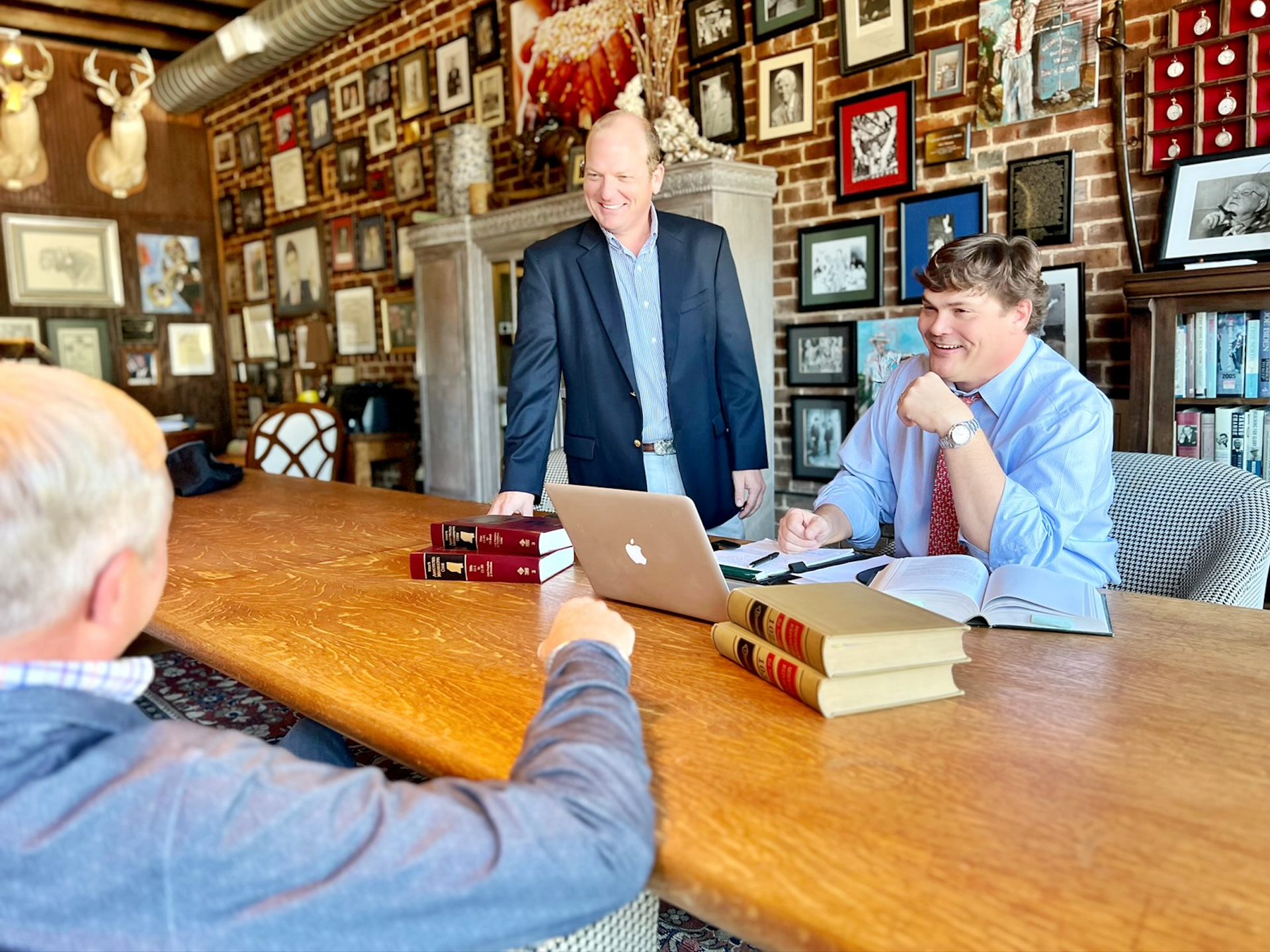 Three men in an office with a long wooden table. Two are standing, one seated, books and a laptop are on the table.