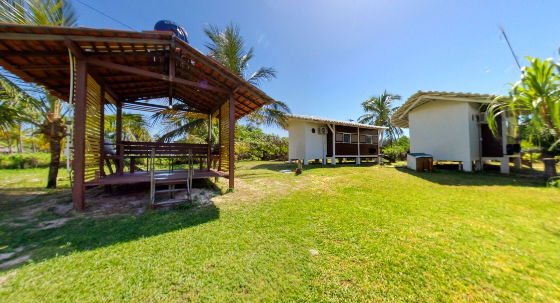 Gazebo de madeira e dois prédios brancos em um gramado verde, céu azul acima.