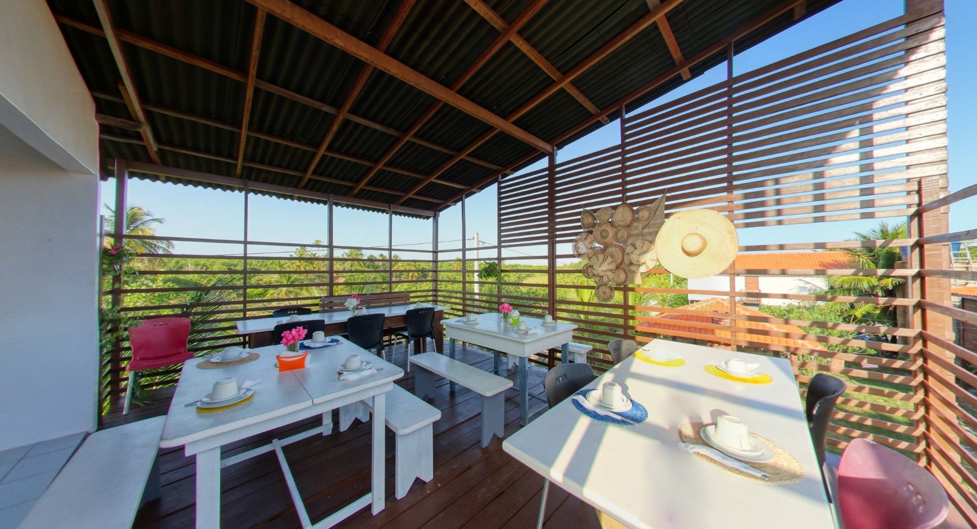 Área de jantar ao ar livre com mesas brancas, bancos e um teto sombreado.