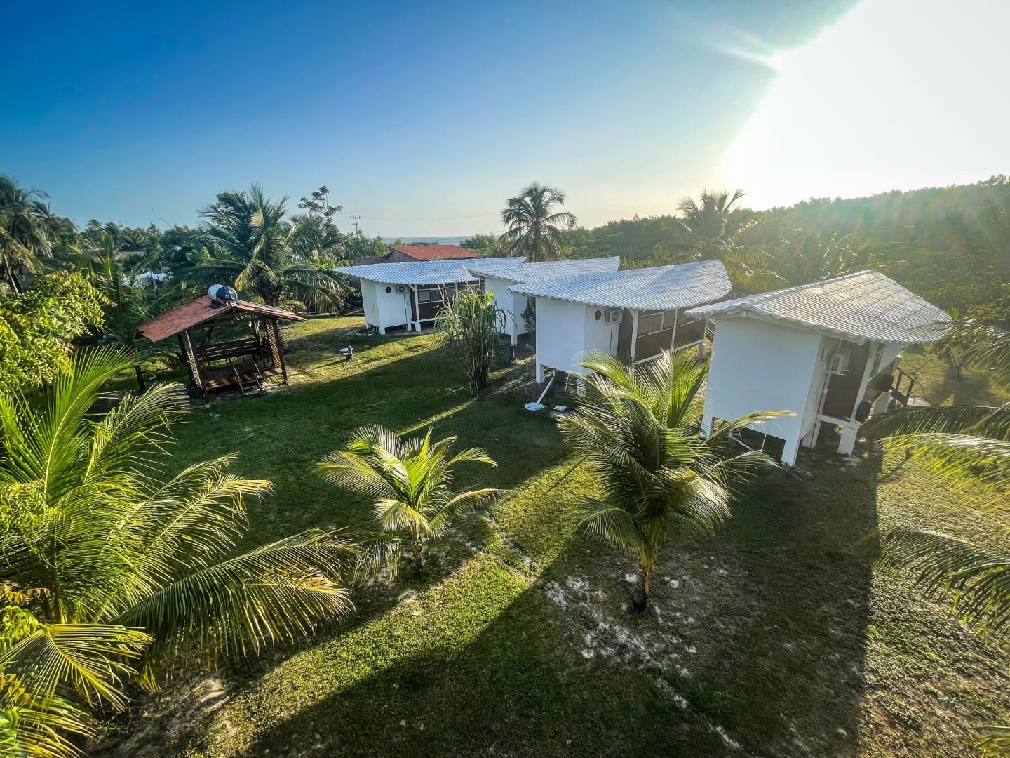 Cabanas brancas com telhados de telha em uma colina gramada, palmeiras, gazebo, luz solar brilhante.