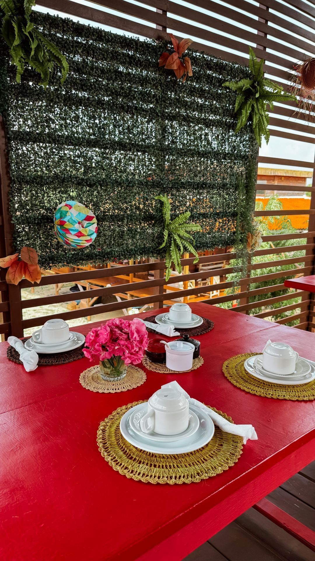 Mesa vermelha posta para chá, com pratos brancos e flores rosas, contra uma parede verde com samambaias.