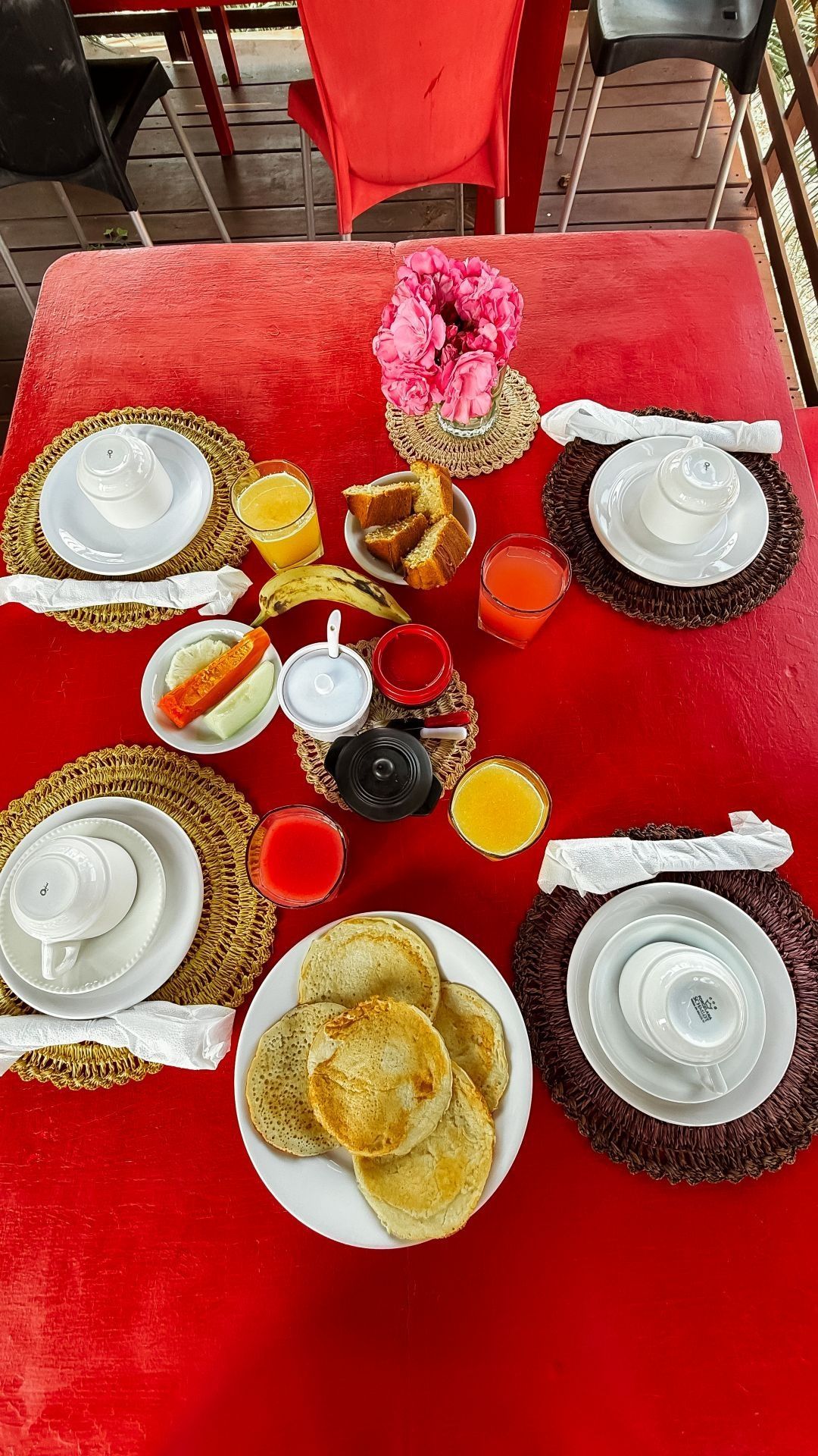 Café da manhã servido em uma mesa vermelha: panquecas, frutas, sucos, xícaras de café e arranjos de flores.
