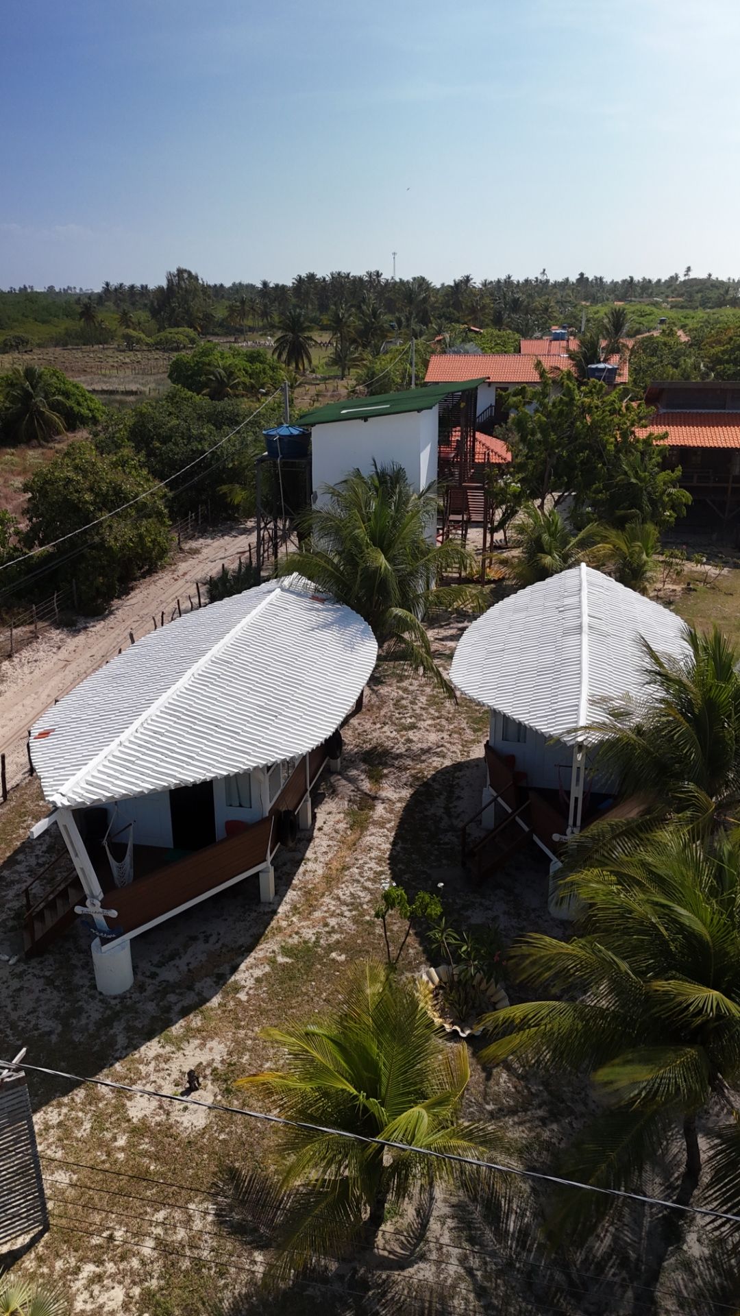 Vista aérea de cabanas à beira-mar com telhados brancos e curvos. Árvores e céu azul ao redor.