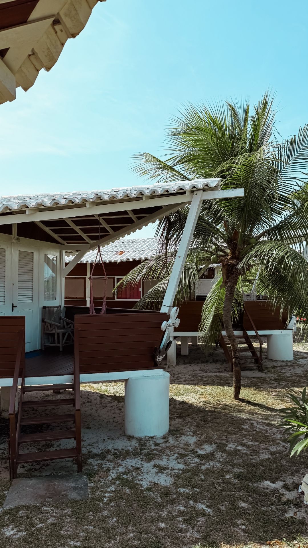 Cabanas brancas à beira-mar sob um céu azul brilhante, com uma palmeira visível.