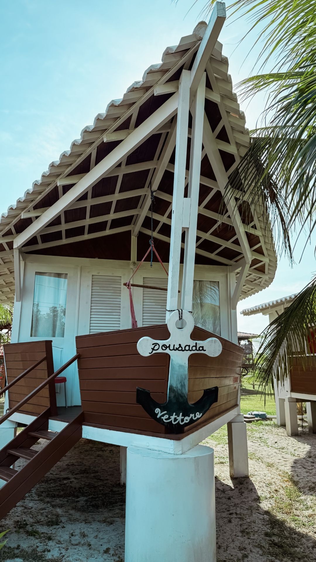 Pequena casa branca e marrom com fachada em forma de barco sobre palafitas, âncora pintada e placa