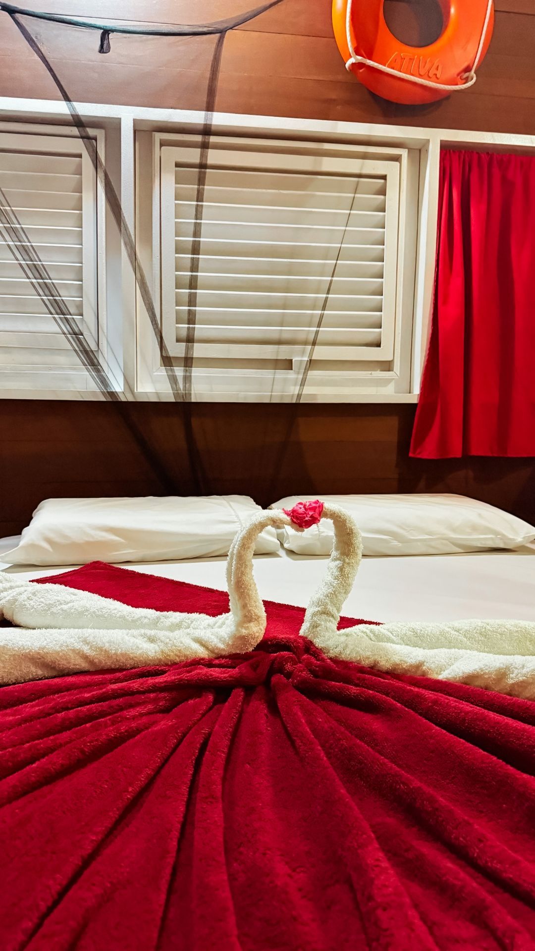 Roupa de cama vermelha e branca com cisnes de toalha; janela e cortinas vermelhas; bóia salva-vidas.