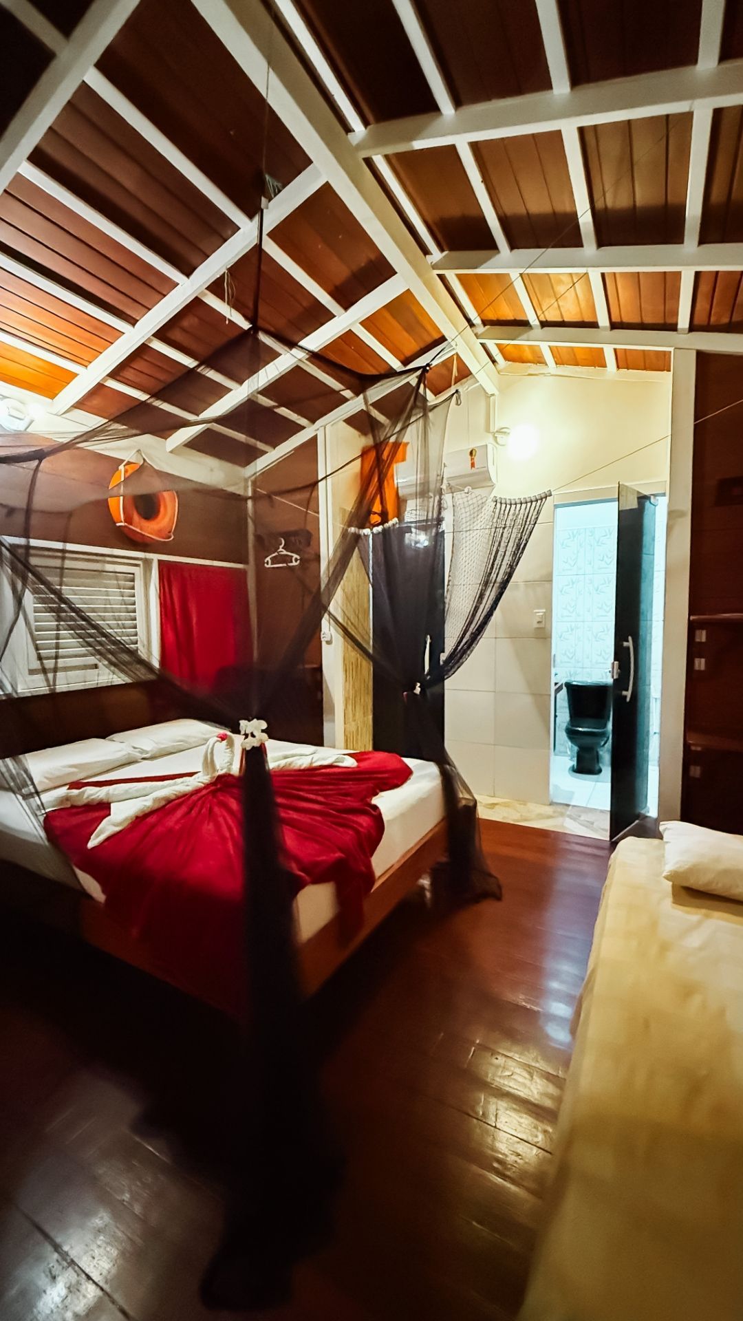 Quarto de madeira com cama, cobertor vermelho, mosquiteiro e banheiro aberto.