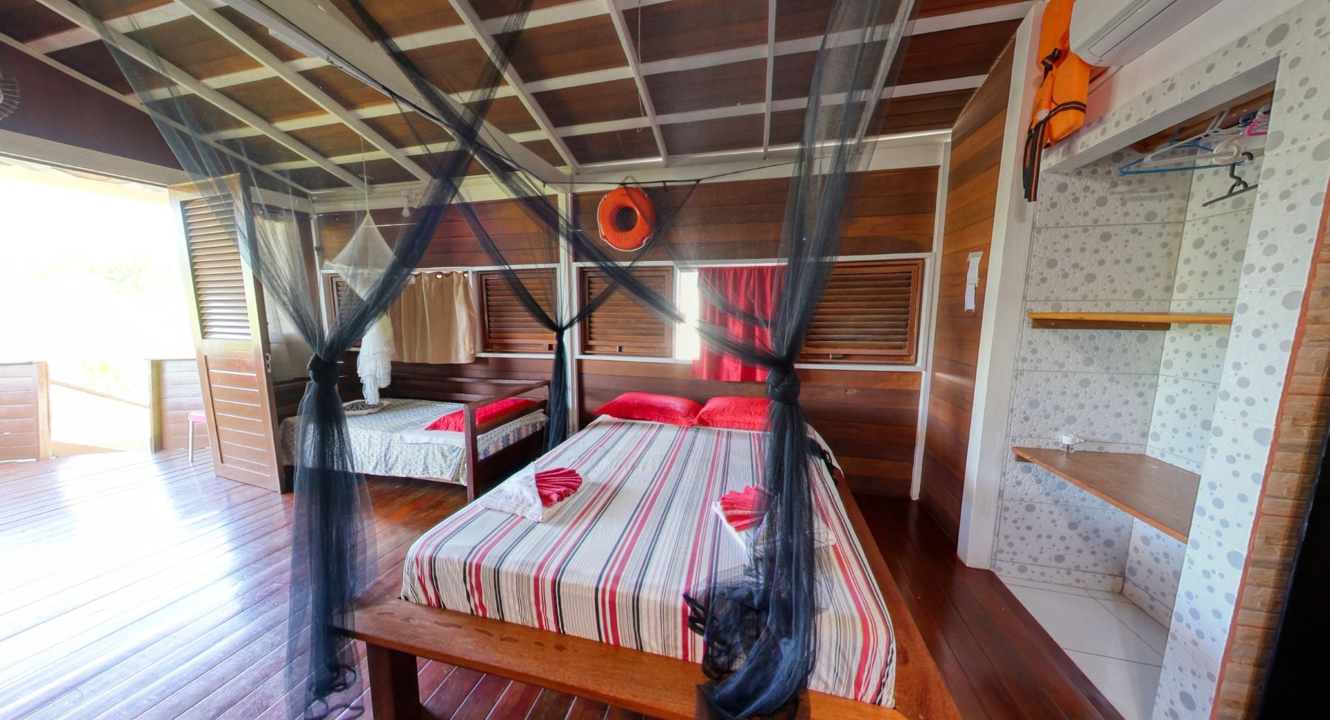 Quarto com duas camas, mosquiteiros, lençóis listrados de vermelho e branco e painéis de madeira.