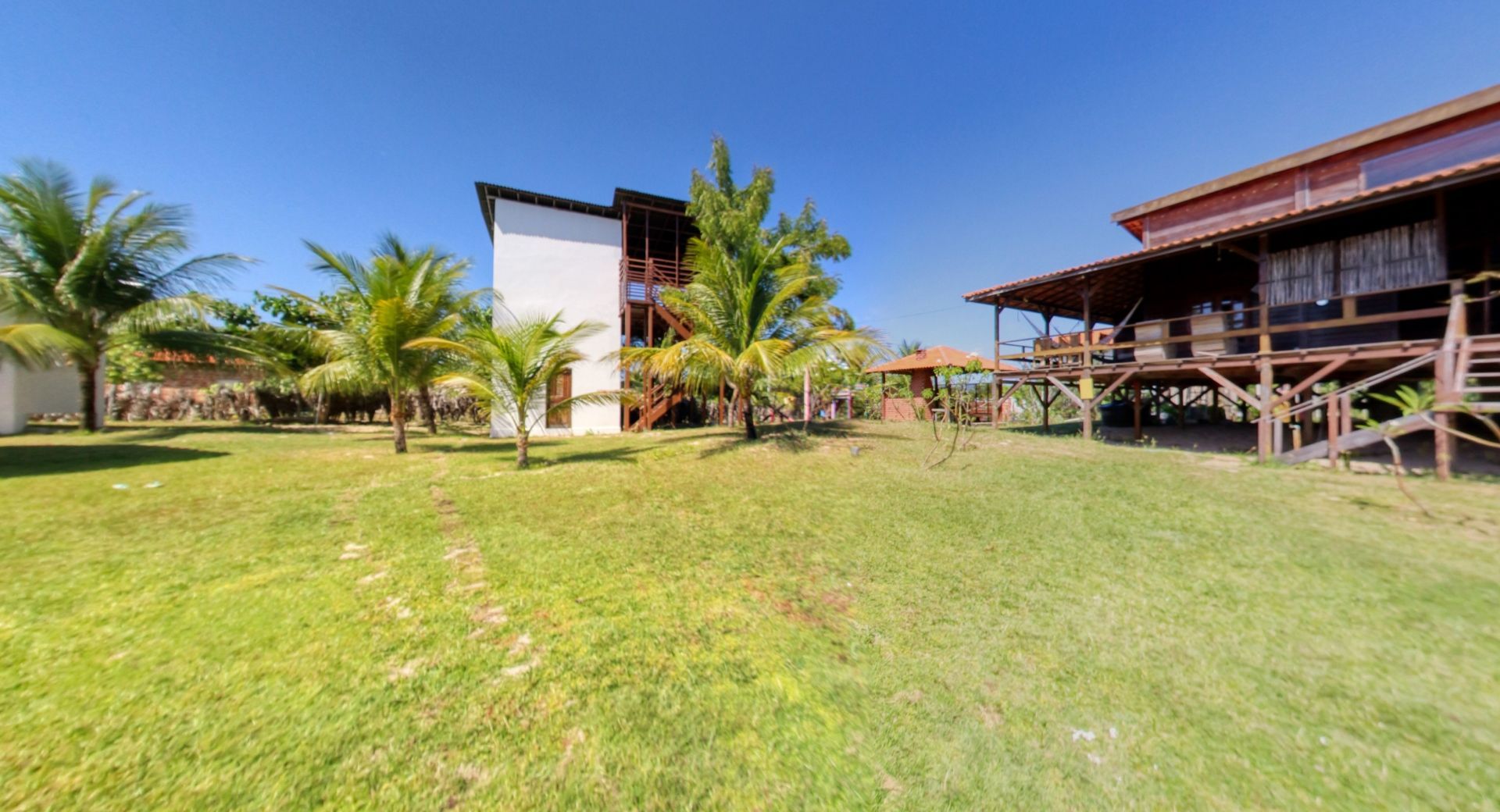 Gramado verdejante com palmeiras e dois edifícios: uma torre branca e uma estrutura de madeira sobre palafitas.