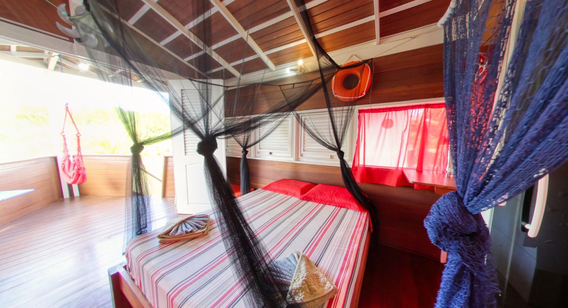 Quarto com mosquiteiro sobre a cama, lençóis listrados de vermelho e branco, paredes e teto de madeira.