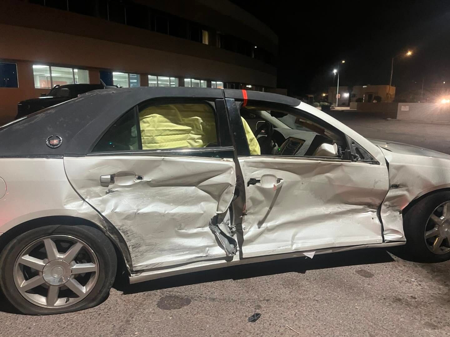 Silver Cadillac sedan heavily damaged on the driver's side, airbags deployed. Scene lit by streetlights at night.