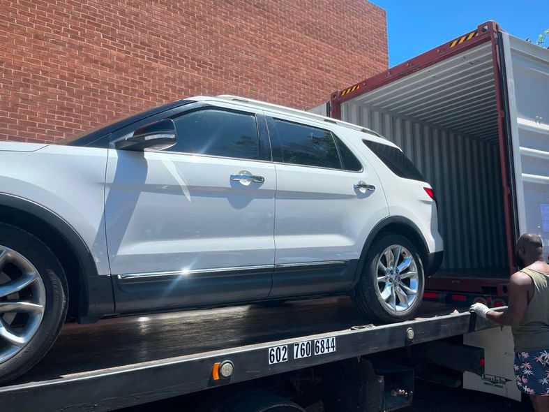 White SUV being loaded onto a flatbed truck and into a shipping container next to a brick building.